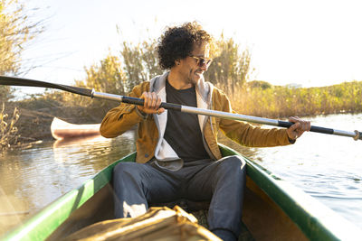 Smiling man wearing sunglasses paddling through oar while sitting in canoe on river