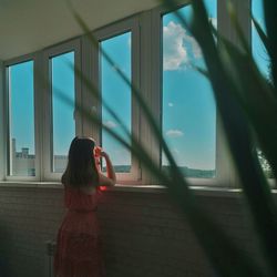 Side view of young woman looking through window while standing in room