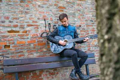 Young man playing guitar while sitting on bench against brick wall