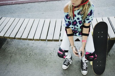 High angle view of girl with skateboard holding mobile phone while sitting on bench at park