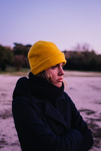 Young woman in hat looking away while standing against sky during winter