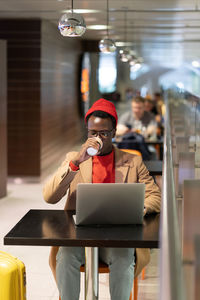 Afro man hipster sitting at cafe in airport terminal drinking coffee or tea works remotely on laptop