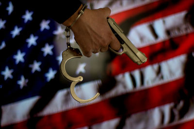 Cropped hand of male criminal holding gun against american flag