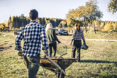 Rear view of male farmer pushing wheelbarrow while following friends at farm