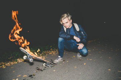 Portrait of young man crouching on road with burning skateboard at night