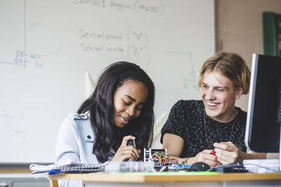 Smiling female student preparing science project with young male friend on desk in classroom at high school