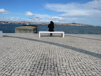 Rear view of woman sitting on bench at cobbled promenade against sky