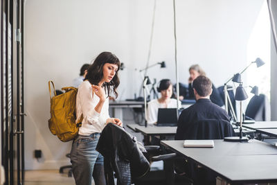 Creative businesswoman with backpack standing by chair in office