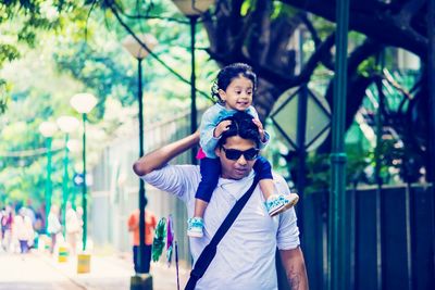 Man carrying daughter on shoulders while walking on footpath