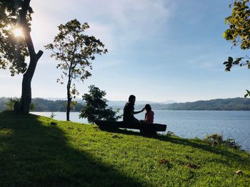 Mother and daughter sitting at lakeshore against sky