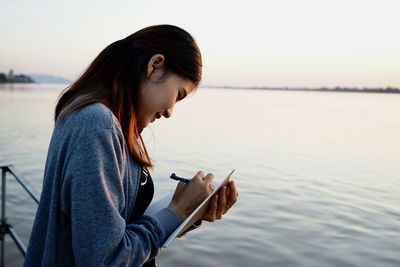 Side view of young woman writing in book by lake against clear sky
