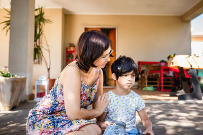 Mother talking to daughter sitting in front of the house watching magazine