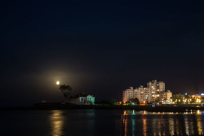 Illuminated buildings by sea against sky at night