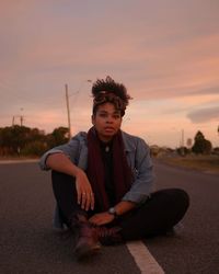 Young woman sitting on road against sky during sunset