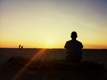 Rear view of silhouette young man sitting on beach against sky during sunset
