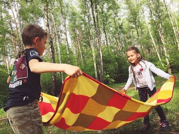 Siblings holding picnic blanket on field against trees in forest