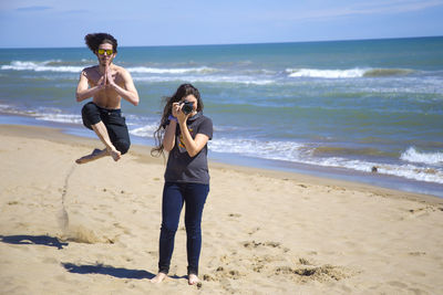 Shirtless man jumping at beach while female friend photographing
