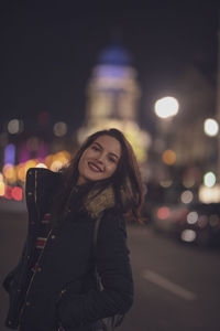 Portrait of young woman smiling while standing on street in illuminated city at night