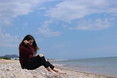 Young woman sitting on shore at beach against sky