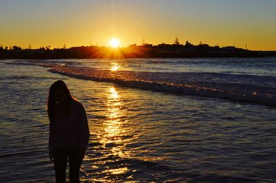 Rear view of silhouette woman standing at beach during sunset