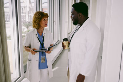 Senior female doctor discussing over medical record with young male colleague while walking in hospital corridor