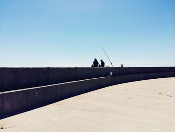 Silhouette men sitting on retaining wall against clear sky during sunny day