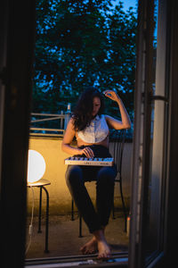Full length of young woman playing piano while sitting on balcony seen through doorway