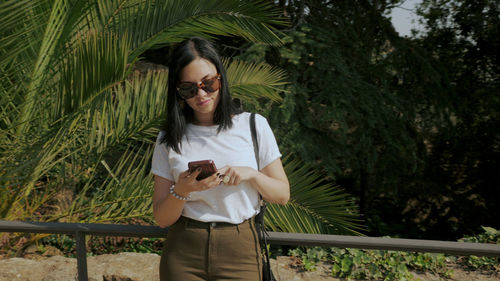 Young woman using mobile phone while standing against palm tree
