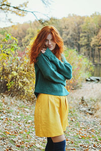 Young redhead woman in green sweater stay near lake in autumn park.