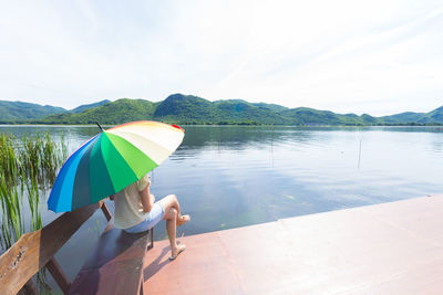 Young woman sitting on bench at jetty over lake against sky