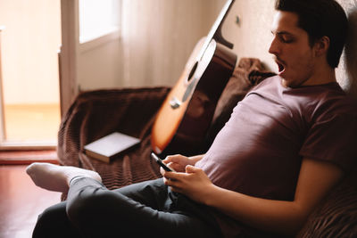 Young man yawning while using smart phone sitting at home