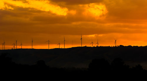 Silhouette wind turbines on mountain against cloudy sky at sunset