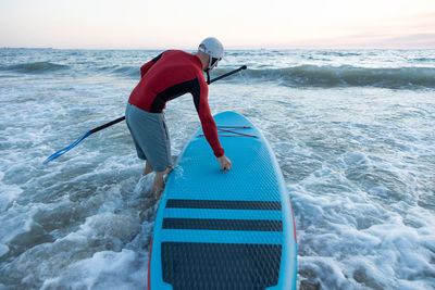 Back view of unrecognizable male surfer in wetsuit and hat carrying paddle board and entering water to surf on seashore