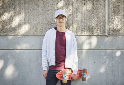 Portrait of young man with skateboard standing against wall in university campus