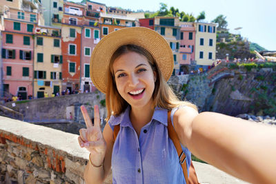 Happy traveler girl taking selfie photo doing victory sign on sunny day in riomaggiore, italy