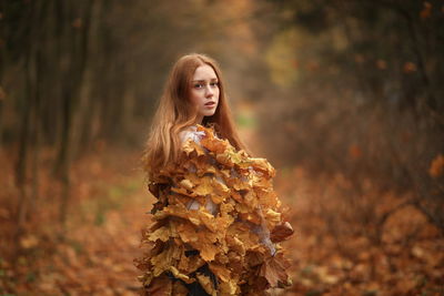 Thoughtful young woman covered with leaves standing at forest during autumn