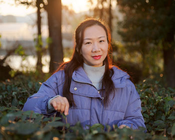 Portrait of smiling mid adult woman in warm clothing standing amidst plants in park during sunset