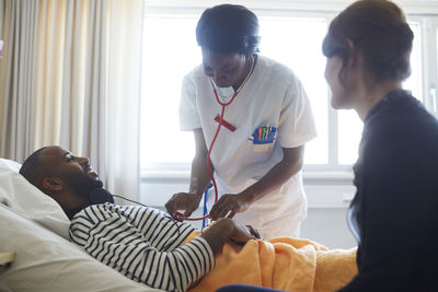 Female doctor examining patient by visitor during routine check up in hospital ward