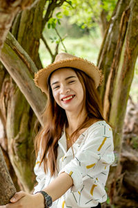 Portrait of happy young woman wearing hat while standing against tree trunks