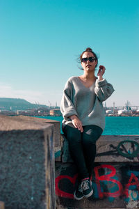 Portrait of young woman sitting on retaining wall against sea