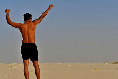 Rear view of shirtless man standing on sand at beach against clear sky
