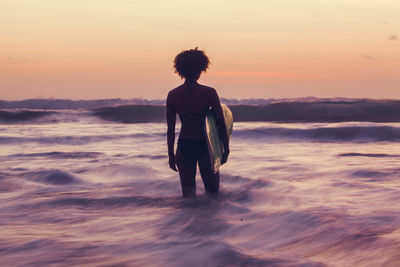 Rear view of woman with surfboard standing in sea during sunset
