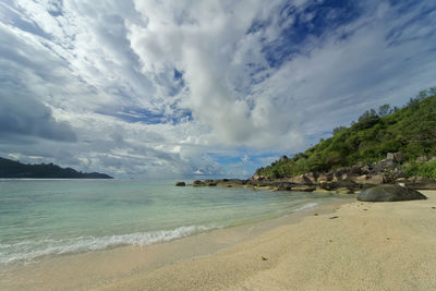 A beach with white and, clear turquoise water, a green coast with boulders going into the water.