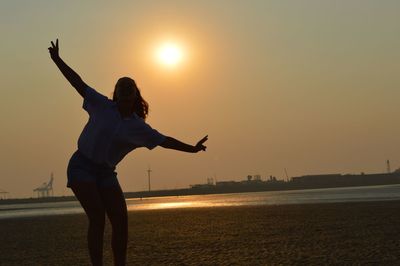 Man with arms outstretched standing on sea shore against sky during sunset