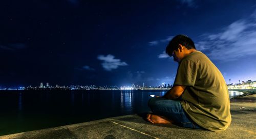 Rear view of man sitting by lake against sky at night