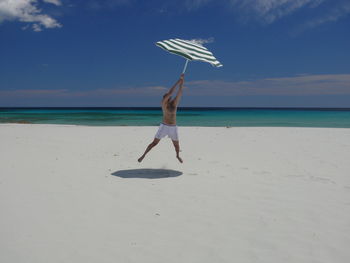 Full length of shirtless man holding umbrella jumping on beach against sky