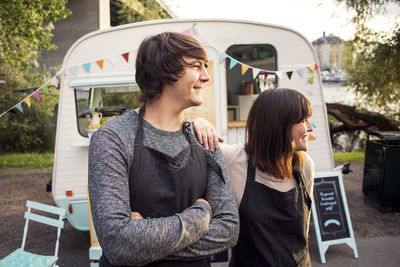 Smiling male and female owners looking away while standing on street against food truck