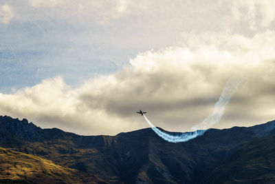 Low angle view of airplane flying above mountains against cloudy sky