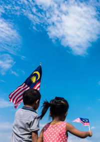 Rear view of friends holding malaysian flags while standing against blue sky