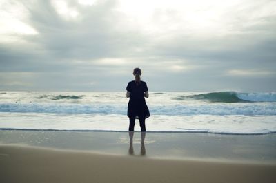 Rear view of man standing at seashore against cloudy sky